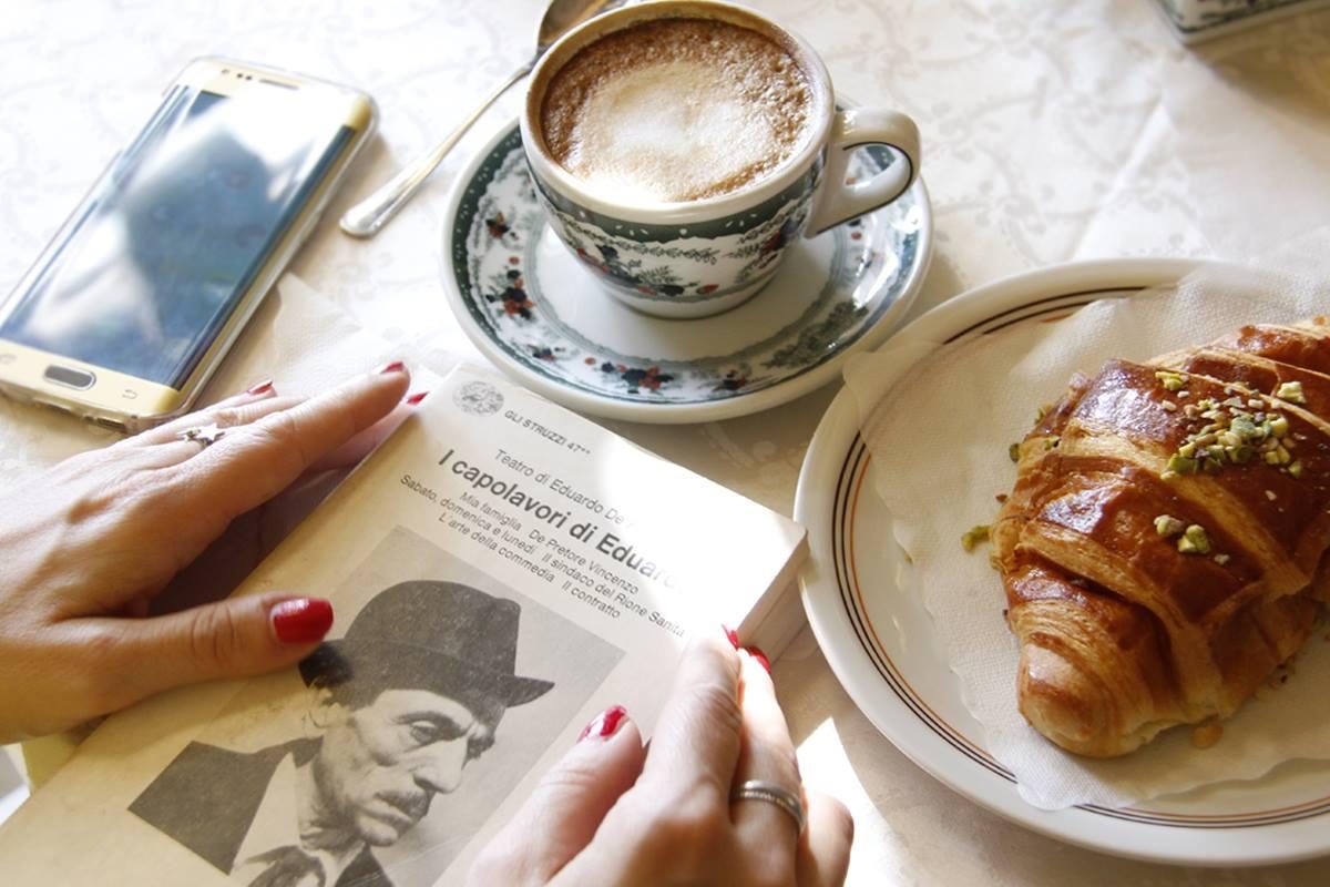 Hände auf einem aufgeschlagenen italienischen Buch, daneben Cappuccino und Cornetto