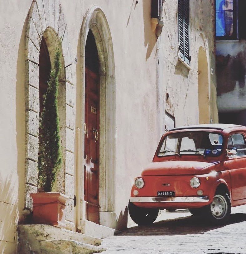 Roter Fiat 500 vor einer Bogentür in Montepulciano, daneben eine Zypresse im Terrakottatopf